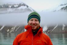 Man in striped wool hat and orange rain jacket stands in front of fjord with mountains in fog