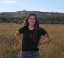 Rikki Held (woman with fair skin and long brown hair wearing a black t-shirt and blue jeans) smiles at the camera in front of a bedrock outcrop with horses in the foreground.