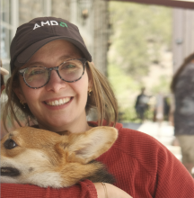 Robyn Marowitz (woman with fair skin, glasses and brown hair pulled back into a ponytail, wearing a hat and a red top, and holding a corgi) smiles at the camera