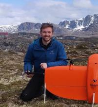 Frederick Hass (man with pale skin, brown hair and a brown beard) smiles at the camera in front of Greenlandic mountains, holding an orange drone in the foreground
